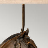 A close-up view of the Eclipse Table Lamp With Tapered Square Shade - Bronze Patina features a horse bust base with a bronze patina. A metal rod extends from the top of the horse's head to support the beige lampshade above, contrasting against the plain light gray background.