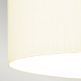 A close-up view of the Fletcher 22" Flush features its cylindrical faux silk ivory shade. The texture of the fabric is visible as the light within gently illuminates the interior, casting a soft glow. Paired with an aged brass fixture, it stands gracefully against a neutral gray backdrop.