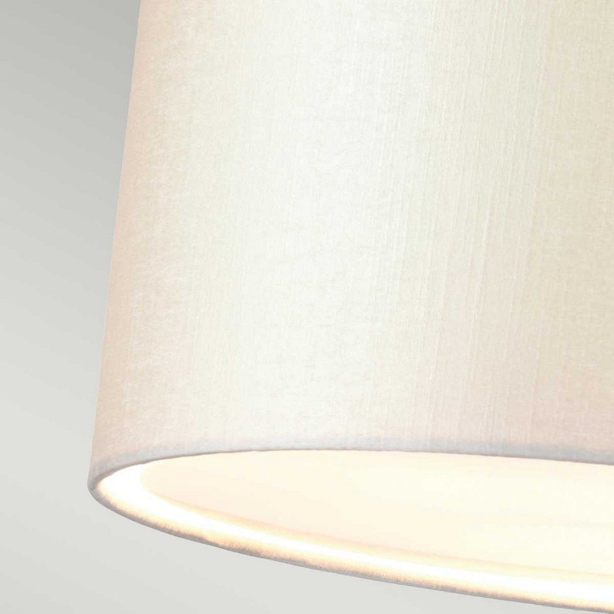 Close-up of an ivory fabric lampshade emitting a soft, warm light set against a light gray background, reminiscent of the elegant Fletcher 34" Flush - Aged Brass with Ivory Shade. The texture of the fabric appears smooth and slightly woven, complementing the aged brass ceiling fixture beautifully.
