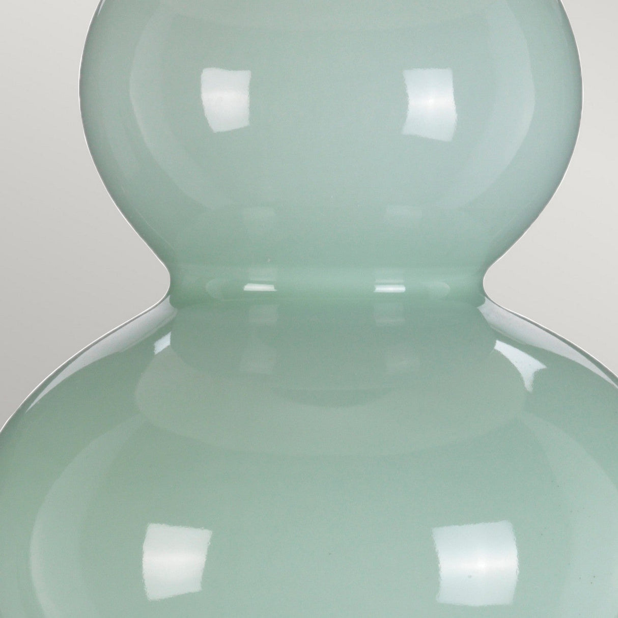 A detailed view of the Harbin Gourd Table Lamp with Tall Empire Shade showcases its smooth, glossy, pastel green surface and distinctive two-tiered shape reminiscent of a gourd. The celadon finish beautifully reflects light, producing bright highlights against the simple light gray background.