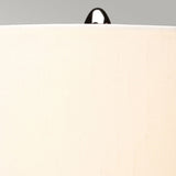 Close-up of a beige lampshade featuring a polished nickel finial atop, set against a neutral gray background. The smooth texture of the lampshade and the reflective quality of the finial beautifully accentuate the elegant style of the Kara Table Lamp - Polished Nickel - Blue Glass.