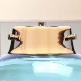 Close-up of the Kara Table Lamp's metallic neck in polished nickel, featuring two adjustment knobs with a reflective gold-like finish. Below is its vibrant blue glass base, and above, a portion of the white lampshade. The background is a neutral light gray.