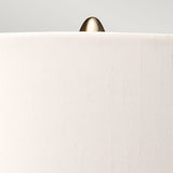 Close-up of a beige fabric lampshade with an aged brass finial on top, featured in the sophisticated Kara Table Lamp - Aged Brass - Dark Green Glass collection, set against a light gray background.