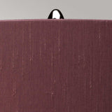 A close-up of a textured, reddish-brown fabric against a gray background reveals a black loop or tag at the top center, suggesting it might be part of the back of a chair or similar object. Envision this sophisticated scene enhanced by the polished nickel and smoke glass base of the Kara Table Lamp - Polished Nickel - Smoke Glass.