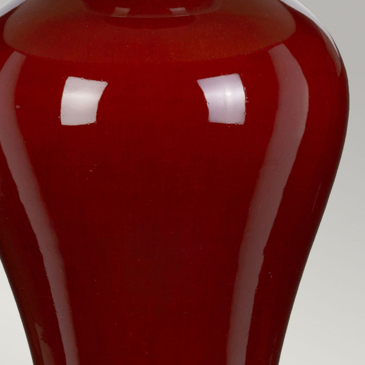 Close-up of a glossy, deep red ceramic vase with a smooth surface and rounded edges, reminiscent of an oxblood finish. The vase reflects light like the Majin Table Lamp With Tall Empire Shade - Oxblood, creating bright spots on its shiny exterior.
