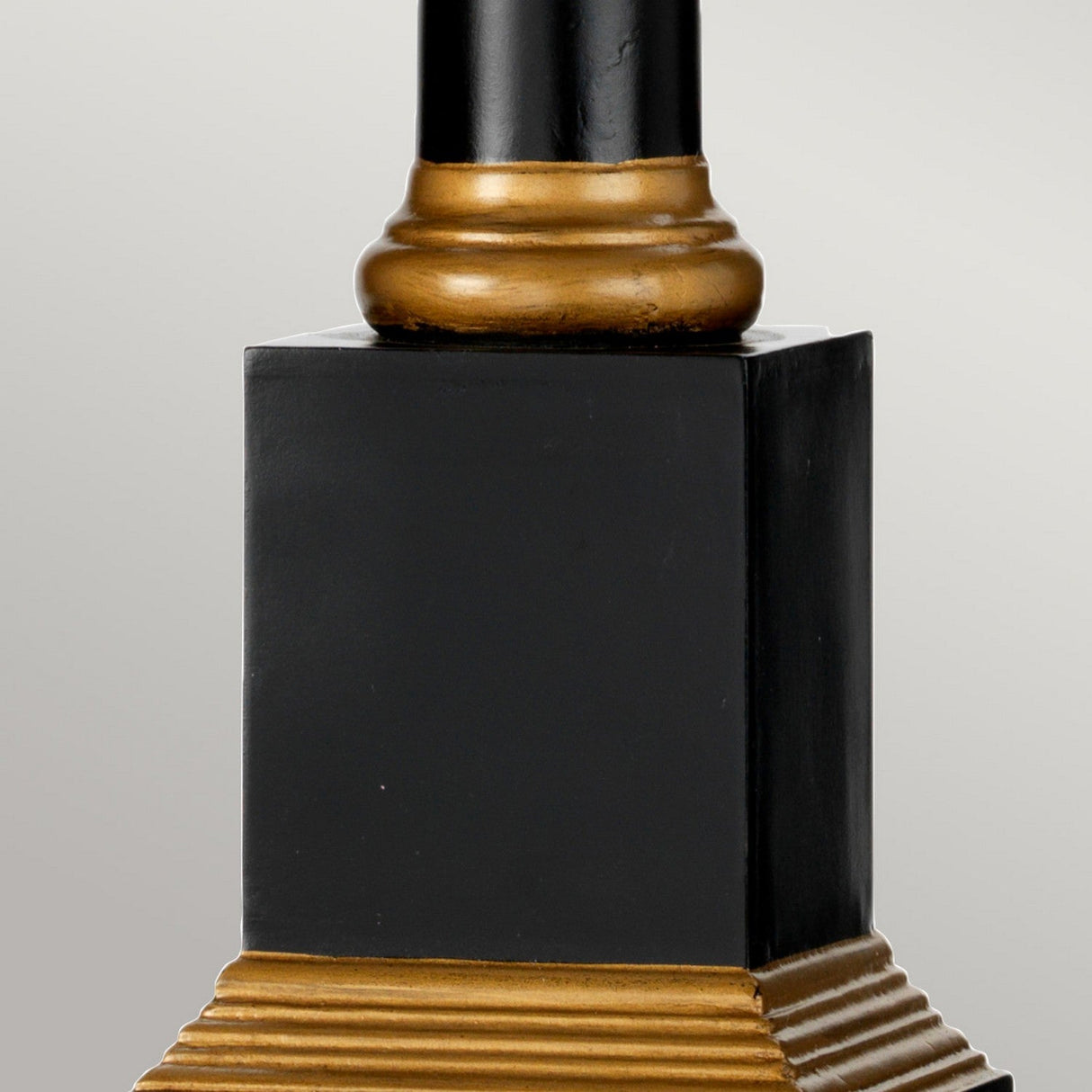 A close-up view of the Peronne Table Lamp With Tall Empire Shade reveals its decorative black column adorned with elegant gold accents. The base showcases a stepped gold design, while a rounded gold ring sits just beneath the main shaft. This striking black and gold motif stands out beautifully against a plain, light grey background.