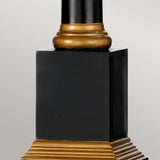 A close-up view of the Peronne Table Lamp With Tall Empire Shade reveals its decorative black column adorned with elegant gold accents. The base showcases a stepped gold design, while a rounded gold ring sits just beneath the main shaft. This striking black and gold motif stands out beautifully against a plain, light grey background.