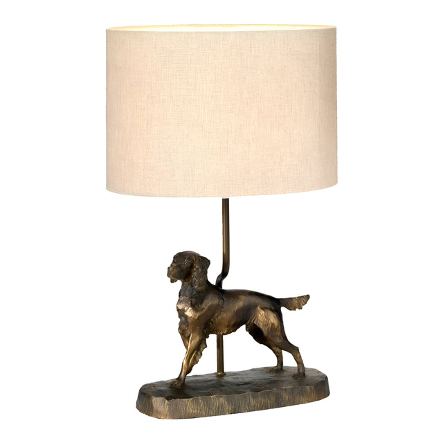 Introducing the Rufus Table Lamp With Oval Shade in an elegant bronze patina, showcasing a base shaped like a dog statuette in a walking pose and completed with a beige cylindrical shade.