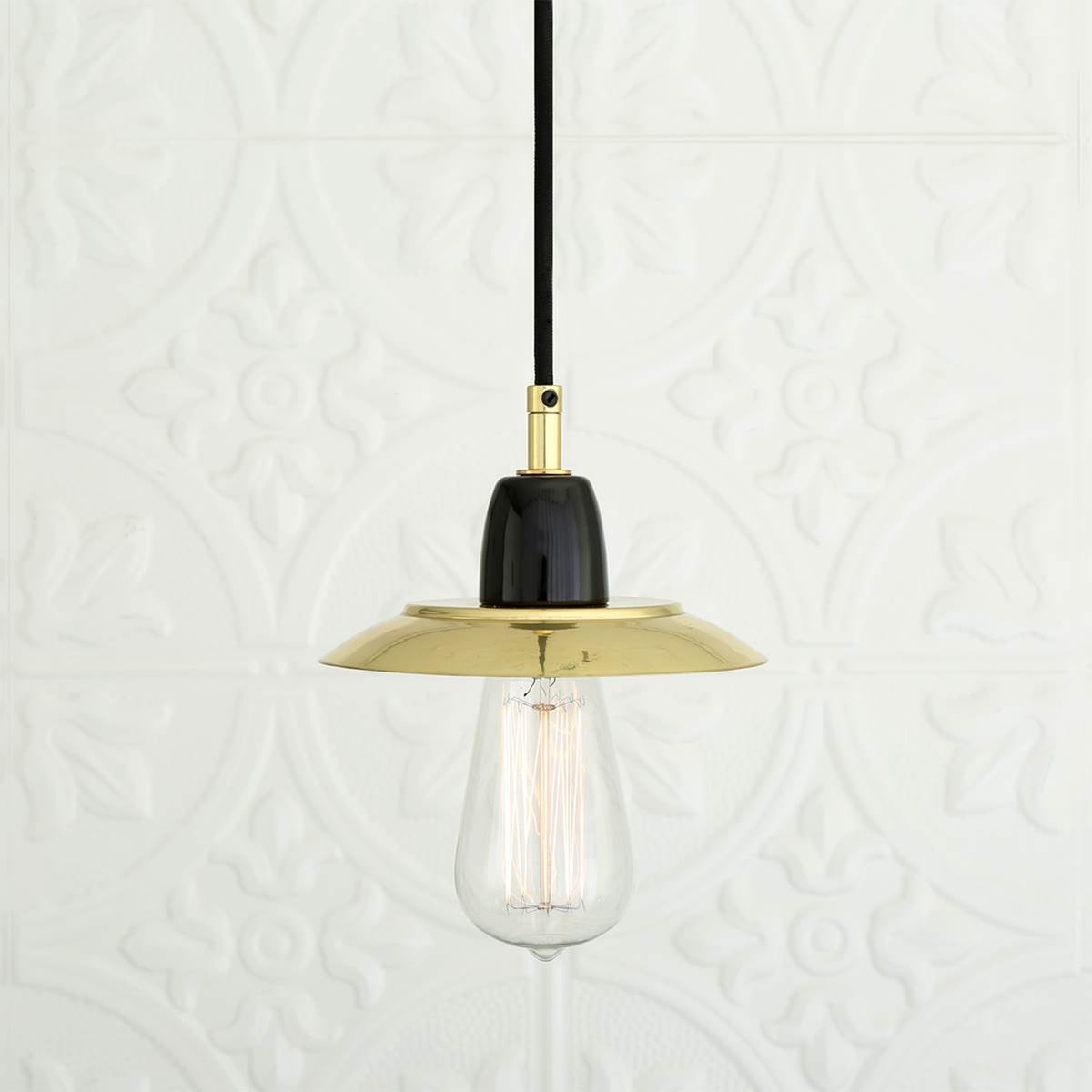 Doon Brass Pendant Light with a shallow, wide-brimmed polished brass shade, black ceramic lampholder, exposed bulb, and black cable, shown against a plain patterned white background.