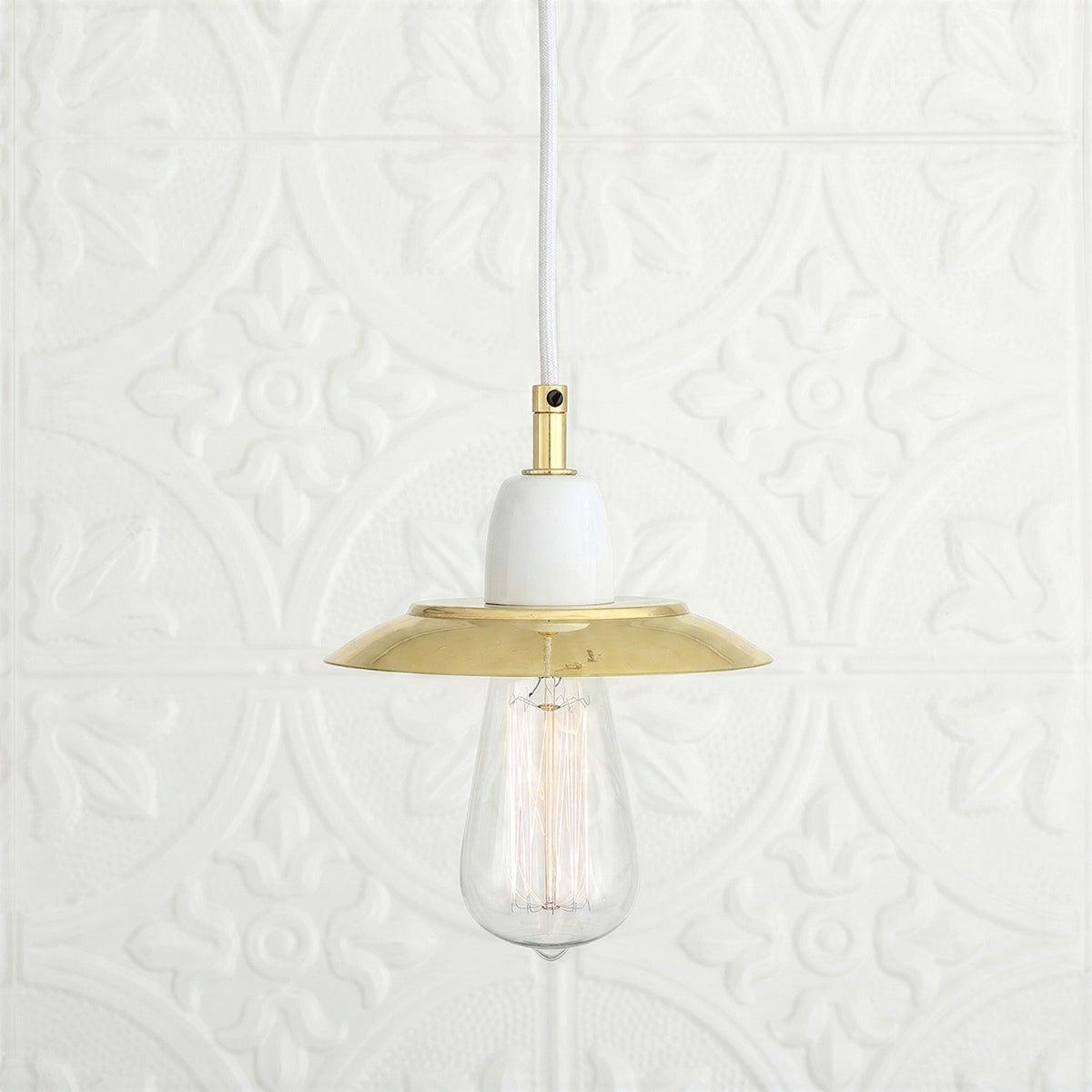 Doon Brass Pendant Light with a shallow wide-brimmed polished brass shade, white ceramic lampholder, white cable, and exposed bulb, shown against a light patterned background.