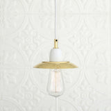 Doon Brass Pendant Light with a shallow wide-brimmed polished brass shade, white ceramic lampholder, white cable, and exposed bulb, shown against a light patterned background.