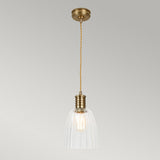 The Douille 1 Light Pendant in aged brass features a vintage design with a twisted cord and ribbed, translucent glass shade that hangs from the ceiling. It casts a warm glow against neutral surroundings, enhanced by an LED filament lamp.