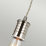 An up-close view of the Douille 1 Light Pendant in polished nickel showcases its industrial, cylindrical design. It hangs from a twisted, fabric-covered cord against a plain, light grey backdrop that accentuates its sleek and modern appeal, beautifully complemented by vintage-style LED filament bulbs.