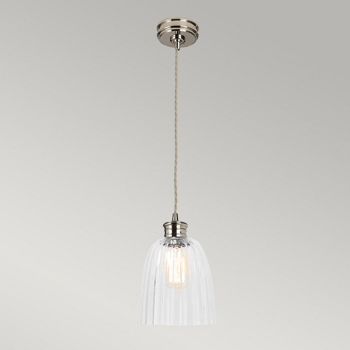 The Douille 1 Light Pendant in polished nickel features a vintage-style industrial design with a clear ribbed glass shade hanging from a twisted cord. The LED filament bulb inside provides a warm glow, perfectly complemented by the fixture's polished nickel base against a light gray background.