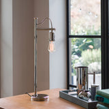 A stylish Douille Table Lamp - Polished Nickel with an exposed vintage-style LED filament bulb is elegantly placed on a wooden desk beside a silver coffee pot and tray, set against the backdrop of a large window featuring a garden view.