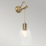 Announcing the Douille Wall Light in aged brass, a wall-mounted fixture that exudes vintage charm and industrial elegance. It features a clear ribbed glass shade and visible bulb, connected by twisted wiring. The light hangs from an elegant bracket with a curved arm, all set against a minimalist background.