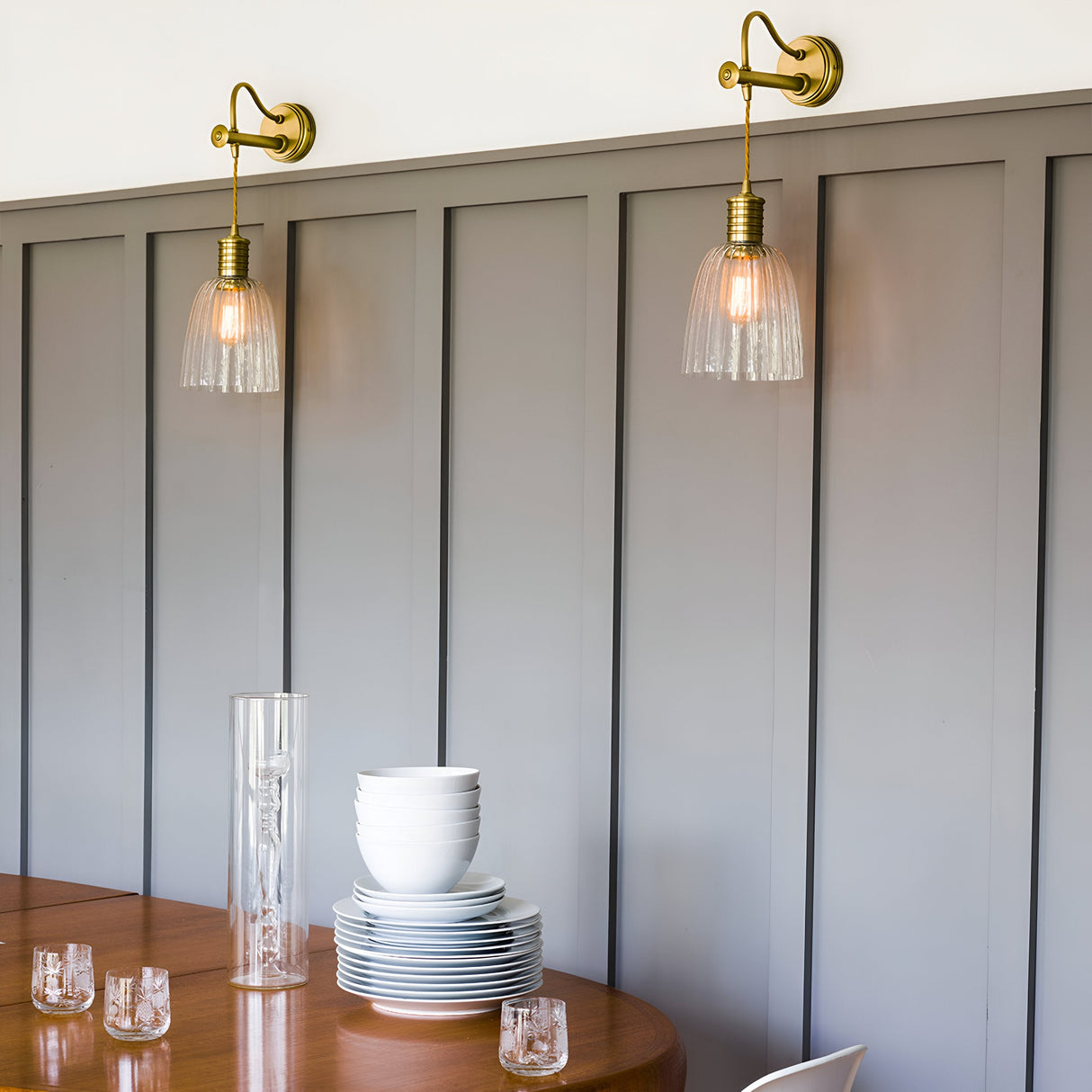 The dining area exudes industrial elegance with Douille Wall Lights in aged brass, featuring fluted glass lampshades on a gray paneled wall. A wooden table below holds stacked white plates, bowls, and crystal glassware, centered by a tall clear vase.