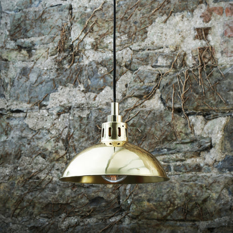 Dovrin Brass Dome Pendant Light IP65, a wide shallow polished brass shade with vented gallery and stepped stem over an exposed bulb, shown against a stone wall.