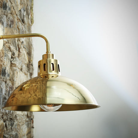 Dovrin Brass Dome Wall Light IP65 in polished brass, showing the deep dome shade, vented collar, and swan-neck arm against a stone wall.