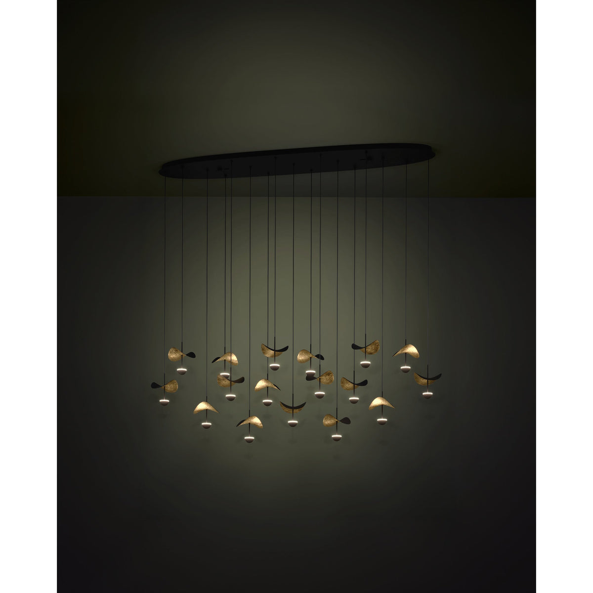 Dracera 17 Light Linear Pendant Light in black steel, with an oval ceiling canopy, fine black cords, and curved leaf-shaped shades in black and gold catching warm LED glow.
