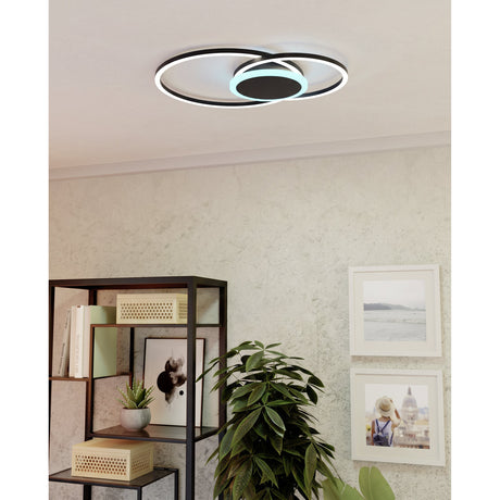 Dralena colour changing LED flush ceiling light in black, two overlapping rings with centre disc, shown mounted on a living room ceiling above a bookshelf and framed wall art.