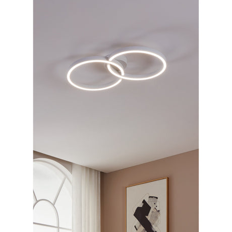 Dralena LED flush ceiling light in white, two interlocking illuminated rings joined at a round hub, shown mounted on a plain ceiling above a hallway with window and framed art.