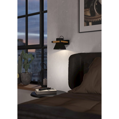 Drasvenne Wall Light in black and brown, with a black conical shade in a square wood frame and black brackets, shown illuminating a bedroom wall beside a headboard.