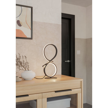 Drelvanne LED Table Lamp in black, a figure-of-eight shaped aluminium and steel design with a glowing white band, shown lit on a wooden sideboard beside a vase and framed art.