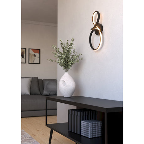 Drelvanne LED Wall Light in black, its ribbon looping into a figure-eight with warm glowing curves, mounted on a light wall above a console table in a living room setting.