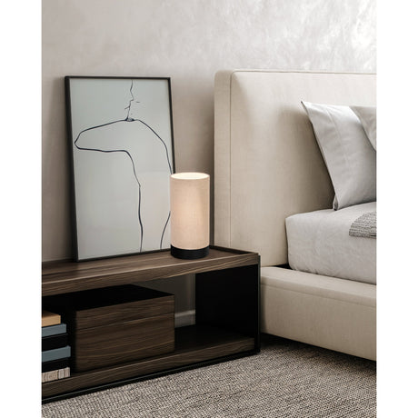 Dremane Table Lamp in black, a cylindrical natural linen shade on a black steel base, shown on a dark wood bedside shelf beside framed line art and a beige upholstered bed.