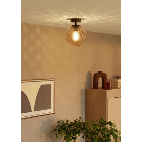 Drilvenne semi-flush ceiling light with dark bronze steel body and dimpled brown glass globe, shown illuminated above a cabinet in a living room setting.
