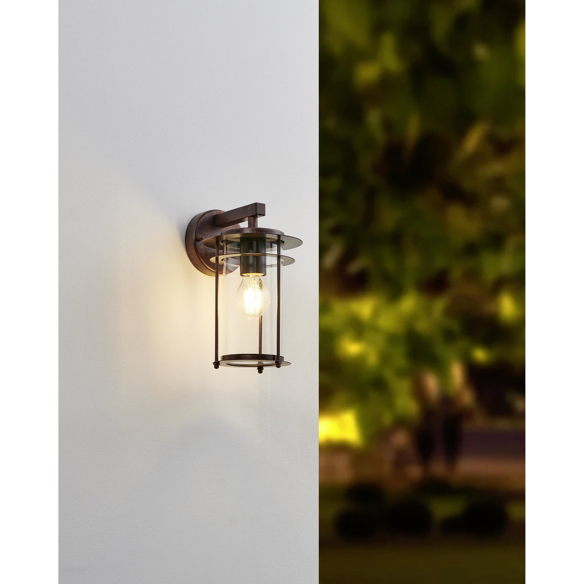 Drimena outdoor wall light in antique copper finish, lantern-style with clear glass cylinder and vertical rods, mounted on a white exterior wall with blurred garden greenery behind.