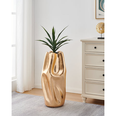 Dunnamore 80cm gold fibreglass plant pot with a crumpled, folded texture and wavy rim, holding a green plant beside a cream chest of drawers in a living room setting.