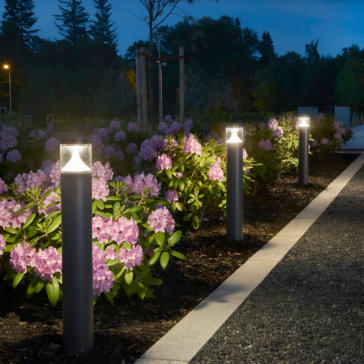 At night, the path is illuminated by energy-efficient Egersund LED Tall Bollard lights in graphite. Purple flowers and greenery line the walkway, with trees visible in the background under a dark blue sky.