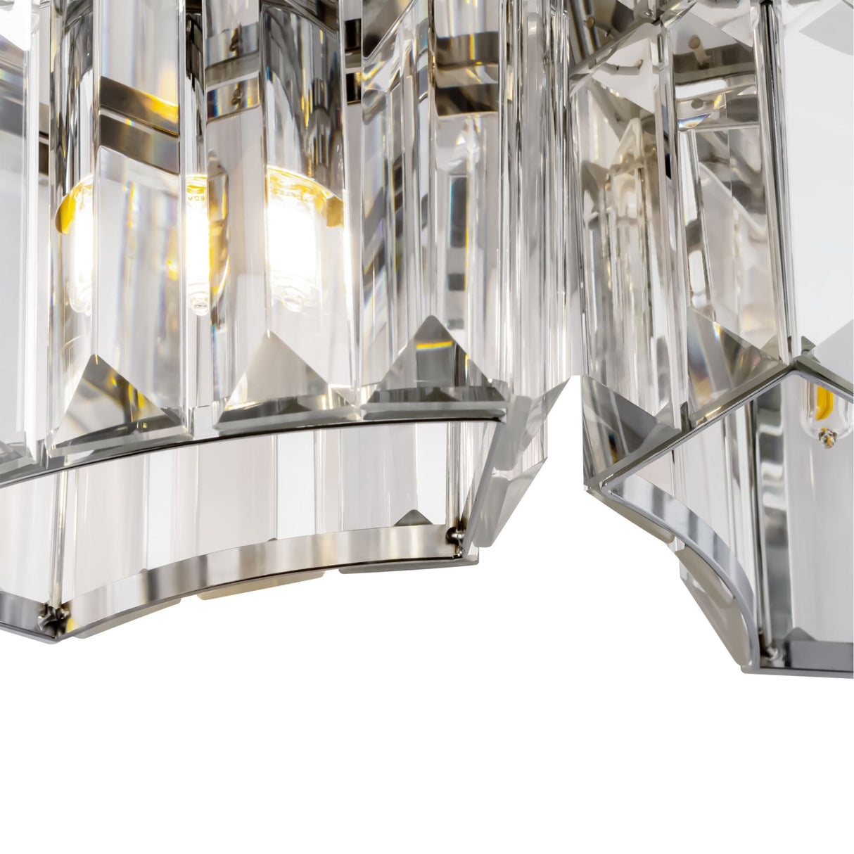 A close-up of the Colline Wall Light - Chrome showcases its geometric, crystal-like glass panels that reflect light like elegant prisms. The sleek design, with multiple faceted pieces, is enhanced by two illuminated bulbs shining through the transparent surfaces.