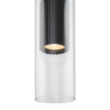 A close-up of the Dynamics Pendant Light showcases a modern cylindrical design with clear glass casing and a ribbed black interior around the bulb. It has a matte black finish, creating a warm, concentrated downward glow.