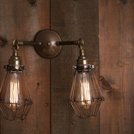 Elsoma Twin Industrial Wall Light in antique brass with a round back plate, two hooked arms, exposed bulbs, and wire cage shades, mounted on a wooden plank wall.