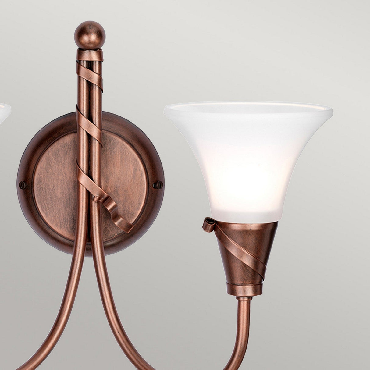 The Emily 2 Light Wall Light in copper patina features a bronze finish and two elegantly curved arms adorned with frosted glass shades. Mounted on a round base, its design includes graceful twists around the arms, creating a classic and sophisticated appearance enhanced by subtle copper patina accents.