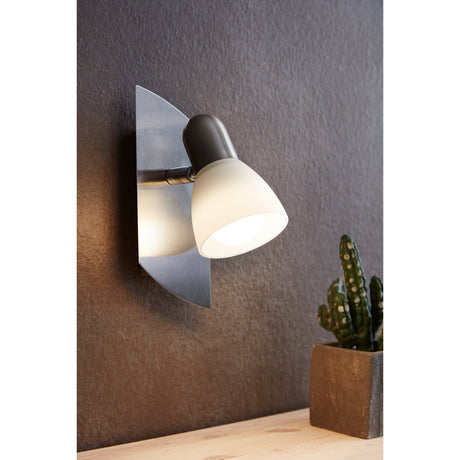 Enea Wall Spotlight in satin nickel with a fan-shaped backplate, swivelling arm, and white satin glass shade, shown illuminated on a dark wall beside a potted cactus.