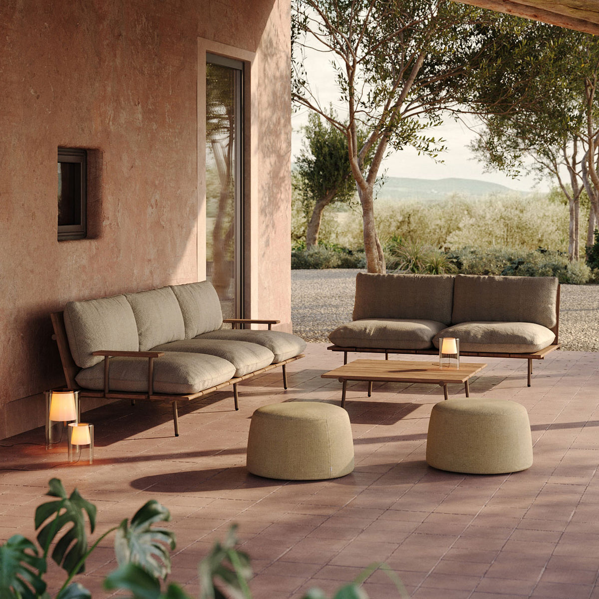 Two round Esen upholstered outdoor pouffes in beige fabric with tapered drum shapes, shown on a terrace beside matching sofas and a wooden coffee table.