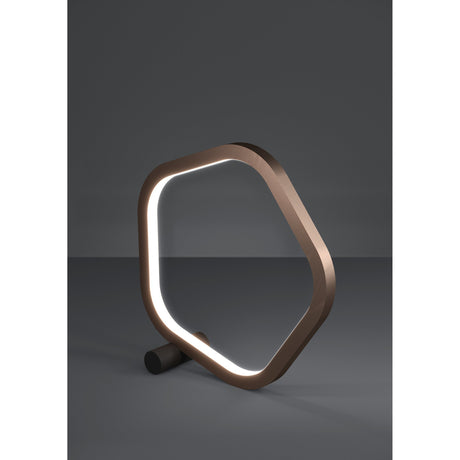 Estrella LED table lamp with a bronze pentagon-shaped ring frame on a black cylindrical foot, illuminated white diffuser, set against a plain grey background.