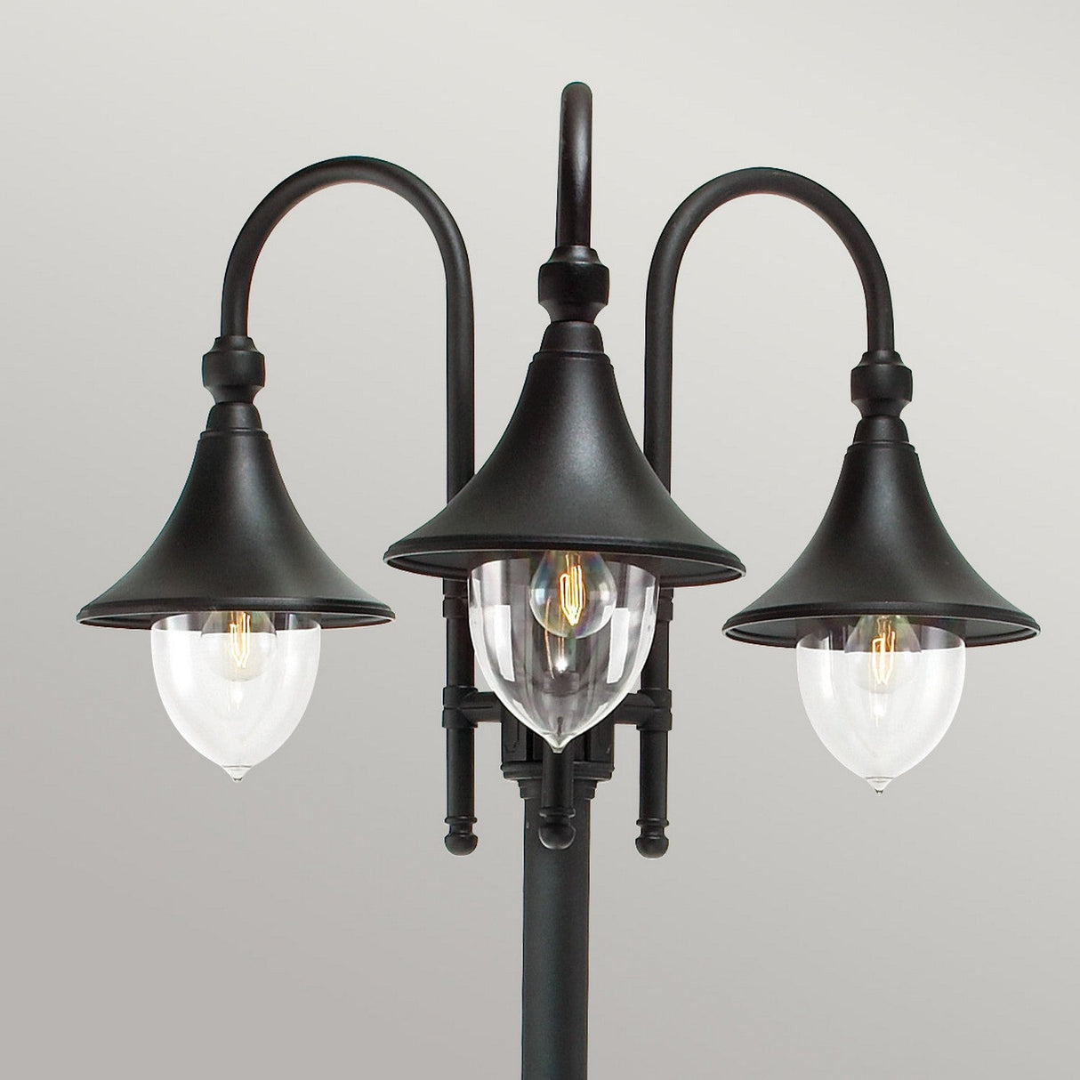 The Firenze 3 Light Lamp Post - Black boasts a sleek black finish and three elegantly curving arms, each supporting a dome-shaped shade. The central transparent shade showcases a filament bulb, with the remaining shades in an opaque design. Ideal for adding charm to any outdoor setting against a simple gray background.