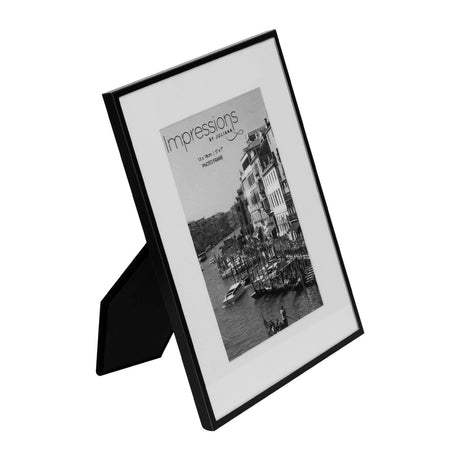 Matt black metal photo frame with white mount, rectangular 7 x 5in design, shown at an angle with fold-out stand, displaying a sample black-and-white cityscape photo on a plain white background.