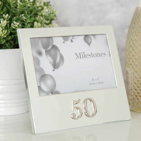 Silver aluminium 6 x 4in photo frame with raised 3D "50" below the landscape window, shown on a table beside a plant in a living room setting.