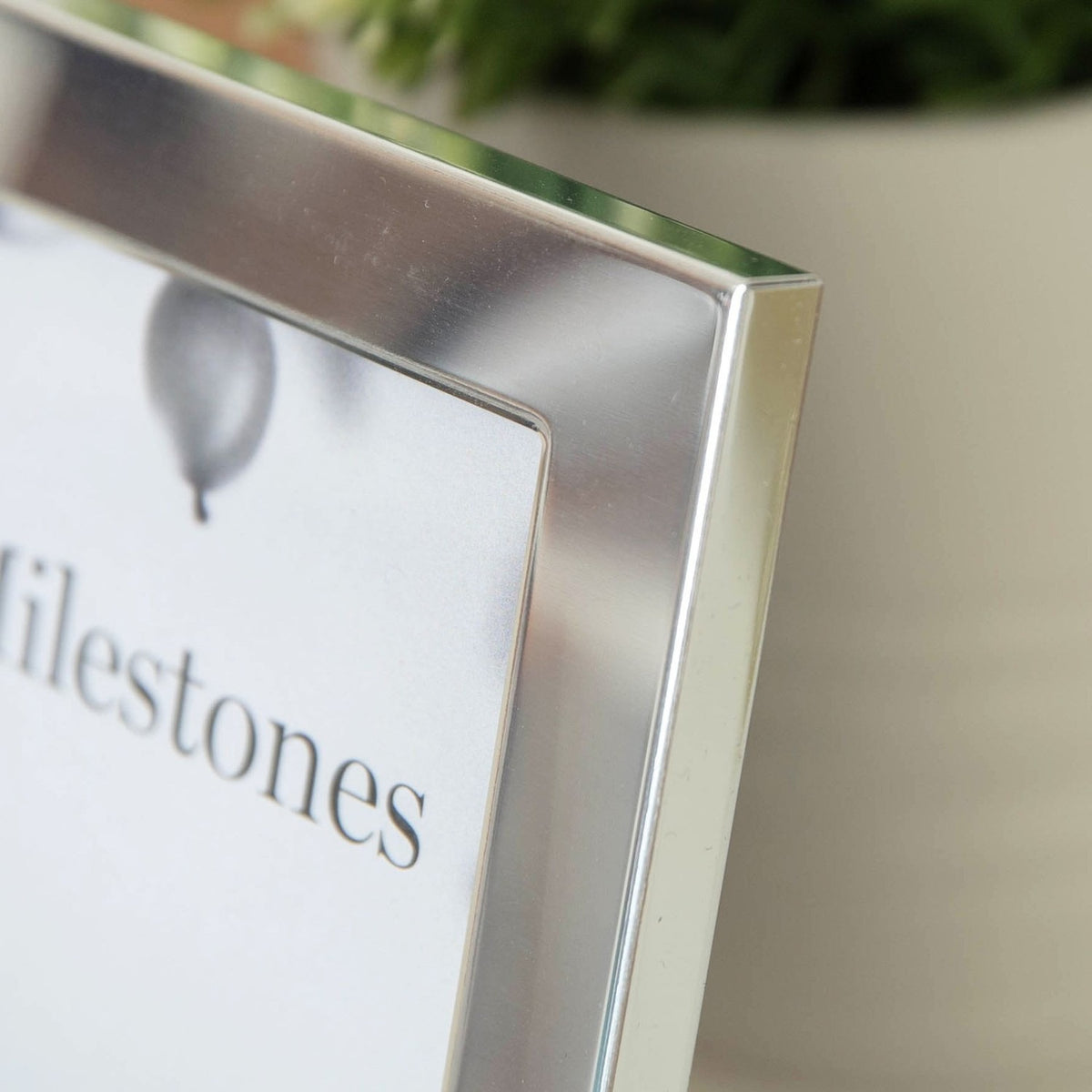 Close-up of the silver aluminium 60th Birthday photo frame's corner, showing the satin metal edge and "Milestones" photo insert, with a blurred plant pot in the background.