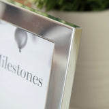 Close-up of the silver aluminium 60th Birthday photo frame's corner, showing the satin metal edge and "Milestones" photo insert, with a blurred plant pot in the background.
