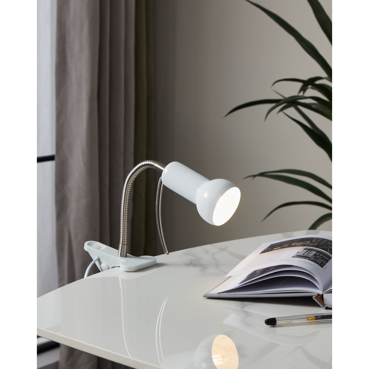 Fabio Clip On Lamp in white and silver, shown clipped to a desk with its flexible gooseneck and bell-shaped shade lit, beside a book in a study setting.