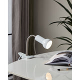Fabio Clip On Lamp in white and silver, shown clipped to a desk with its flexible gooseneck and bell-shaped shade lit, beside a book in a study setting.