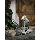 Falmessa Garden Spike Light in stainless steel, with round spotlight head on a slim stem and ground spike, installed among stones and foliage near a doorway.