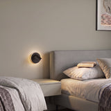 Fanzarotta 1 round LED wall light in black steel with an adjustable cylindrical reading spot, shown glowing beside a bed in a bedroom setting.